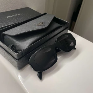 Svarta solglasögon från Prada - Snygga svarta solglasögon från Prada med breda skalmar och cool, kantig form. Kommer med originaletui, putsduk och box. Perfekt accessoar för dig som vill sticka ut med en lyxig och trendig look.