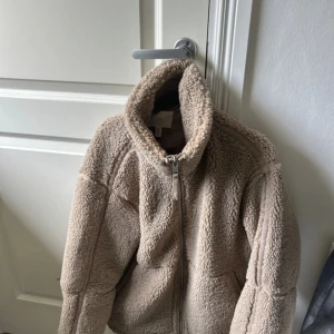 Beige teddyjacka med dragkedja - Mysig beige teddyjacka från H&M med hög krage och hel dragkedja framtill. Jackan har en oversized passform och är tillverkad i fluffigt teddy-material som håller dig varm under kyliga dagar. Perfekt för lager-på-lager och chill vardagsstil.