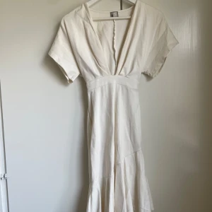 Vit långklänning med v-ringning - Elegant vit långklänning med djup v-ringning och korta ärmar. Klänningen har en markerad midja och ett lätt utsvängt fall nertill. Perfekt för sommarens alla tillfällen och ger en stilren och fräsch look.