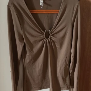 Beige långärmad topp med ringdetalj - Säljer en beige långärmad topp från H&M med djup v-ringning och snygg ringdetalj framtill. Toppen har en tight passform och är tillverkad i mjuk bomullsmix. Perfekt för dig som gillar trendiga och stilrena plagg.