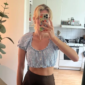Blommig croppad blus från Zara - Ljusblå croppad blus från Zara med vita små blommor och puffiga korta ärmar. Blusen har volangdetaljer vid halsringning och nederkant samt knappar framtill. I nyskick, inga skavanker! 