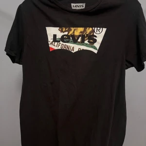 Svart Levis t-shirt med tryck - Svart t-shirt från Levis i bomull med ikoniskt tryck framtill som visar en björn och texten 'California'. Klassisk passform och rund hals. Perfekt för dig som gillar streetstyle och vill ha en cool statement-tee.