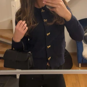 Mörkblå kofta med gulddetaljer - Snygg mörkblå kofta med unika guldfärgade rosettknappar framtill. Koftan har lång ärm och en klassisk, rak passform som passar perfekt till både jeans och kjol. Materialet känns mjukt och är stickat för en extra mysig känsla. endast använd en gång