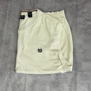 Vit stickad väst från Chaps XXL - Klassisk vit stickad väst från Chaps i storlek XXL. Västen har v-ringning med en diskret rand i brunt vid kragen och broderad logga på bröstet. Tillverkad i mjuk bomull och polyester, perfekt för lager-på-lager-stil.