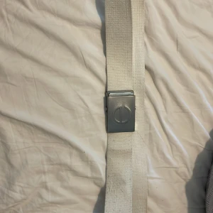 Vitt tygbälte med metallspänne - Snyggt vitt bälte i kraftigt tyg med ett stilrent metallspänne. Spännet har en enkel design med en cirkelformad detalj. Perfekt för att ge en clean och modern touch till din outfit.