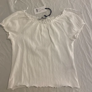Vit offshoulder blus XS - Superfin vit blus från Lager 157 i offshoulder-modell med korta ärmar och lätt rynkad struktur. Toppens material känns luftigt och perfekt för varma dagar. Söt volangkant nertill och vid ärmslut ger en extra touch. Passar dig som gillar en enkel men trendig look. Den är aldrig använd och lappen sitter fortfarande kvar. 