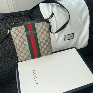 Gucci axelväska med GG-mönster - Säljer en snygg axelväska från Gucci med klassiskt GG-mönster i beige och grått. Väskan har gröna och röda ränder i mitten, svarta skinn-detaljer och dragkedja upptill. Perfekt storlek för det viktigaste och riktigt ikonisk design. Kvitto finns!