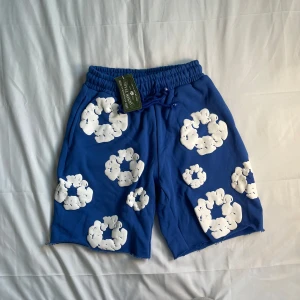 Denim Tears Shorts - 🔥 Denim Tears Wreath Shorts – Blå (Storlek M) 📦 Helt nya med etiketter |  Ett enastående par med Denim Tears ikoniska vita bomullskransgrafik på en djärv blå bas. Kraftig bomull, råa fållar och perfekt för sommarens passform.  🧵 Resår i midjan + dragsko 🧼 Aldrig använda – etiketterna sitter kvar 🌀 Sällsynta och slutsålda  📩 Öppen för erbjudanden och paket – skicka ett direktmeddelande