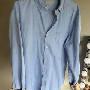 Ljusblå skjorta från Burberry Brit - Snygg ljusblå skjorta från Burberry Brit i 100% bomull. Klassisk button-down krage, bröstficka med diskret broderad logga och ikoniskt rutigt tyg på insidan av kragen och manschetterna. Perfekt för dig som gillar stilrena och tidlösa plagg.