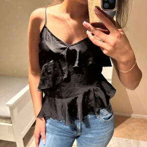 Svart volangig topp i satin - Säljer en svart topp med volanger och en sidsläp. Toppen är från Gina Tricot. Använd en/två gånger.