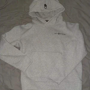 Ljusgrå hoodie från Sail Racing - Snygg ljusgrå hoodie från Sail Racing med svart logga på bröstet och tryck på huvan. Ficka framtill och mjukt material som känns skönt mot huden. Perfekt för dig som gillar en stilren och sportig look.