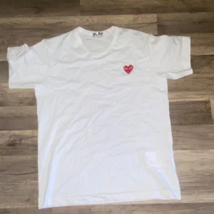 Vit Comme des Garçons Play t-shirt - Vit t-shirt från Comme des Garçons Play med det ikoniska röda hjärtat med ögon broderat på bröstet. Klassisk rund halsringning och korta ärmar. Tillverkad i mjuk bomull som känns skön mot huden. Perfekt för en clean och stilren look.