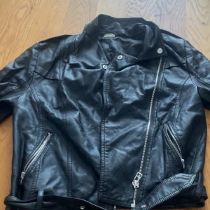 Svart skinnjacka med dragkedjor, C, 40 - Snygg svart skinnjacka från C i storlek 40. Jackan har coola dragkedjor framtill, snedställda fickor och bälte i midjan. Klassisk bikerstil med lång ärm och detaljer i silver. Perfekt för dig som vill ha en edgy look.