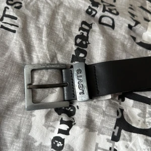 Svart skärp i skinn från Levi's - Snyggt svart bälte från Levi's i 100% skinn med klassiskt silverfärgat spänne och präglad logga vid fästet. Stilren och enkel design som passar till jeans eller chinos. Bältet är rakt och har flera hål för justering.