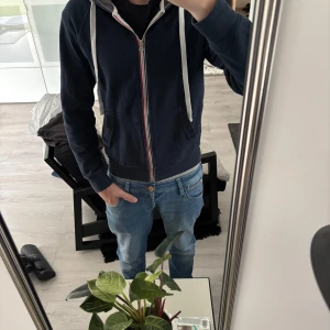 Mörkblå hoodie med dragkedja  - Mörkblå hoodie med vit snörning och dragkedja framtill. Tröjan har huva, kängurufickor och en rödvit detalj längs dragkedjan. Perfekt för chill dagar och enkel att matcha med jeans eller joggers.