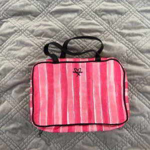 Randig rosa necessär från Victoria's Secret - Superfin necessär från Victoria's Secret i rosa och vita ränder. Praktisk modell med flera genomskinliga fack och dragkedjor, samt en krok för upphängning. Perfekt för att organisera smink eller hudvård när du är på språng.