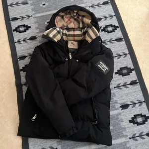 Svart pufferjacka från Burberry S - Svart pufferjacka från Burberry med klassiskt rutigt foder i huvan. Jackan har dragkedja framtill, två fickor med dragkedja och en patch med Burberry London England på ärmen. Perfekt för kalla dagar och riktigt snygg streetstil. Och den har en håll i insidan men de syns inte utanför 