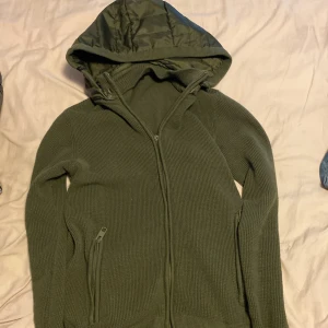 Indicode cardigan - Säljer en olivgrön stickad hoodie med dragkedja och två fickor framtill. Väldigt sällsynt har varigt slutsåld jätte länge. Sitter bra på mig som är 170. Dragkejds grejen är trasig men den funkar lika bra endå