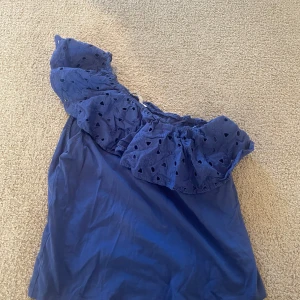 Blå offshoulder topp från MNG - Supersöt marinblå offshoulder topp från mango med volang och broderade detaljer upptill. Toppen är ärmlös och har en luftig känsla, perfekt för varma dagar. Snyggt broderat mönster på volangen ger en extra touch.Tröjan är i bra skick och priset går att diskuteras.Tröjan är ganska liten i storlek skulle jag säga!!!