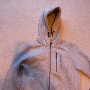 Grå hoodiejacka från Sail Racing - Grå hoodiejacka från Sail Racing med dragkedja framtill, bröstficka med dragkedja och broderad logga på bröstet. Jackan har justerbar huva med snörning och är tillverkad i mjuk bomullsmix. Perfekt för chill dagar eller när du vill ha en sportig look. Finns ett litet slitage på armen men märks ej på!