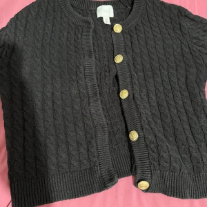 Svart kabelstickad kofta från H&M - Svart kabelstickad kofta från H&M med snygga guldknappar framtill. Koftan har rund halsringning, ribbade muddar och en croppad passform. Perfekt för lager-på-lager och ger en klassisk vibe till din outfit.