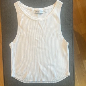 Vit ribbad linnetopp från Zara - Säljer en fräsch vit ribbad linnetopp från Zara i storlek M. Toppen är ärmlös med klassisk rund halsringning och har en tight passform. Perfekt till jeans eller kjol för en clean och enkel look.