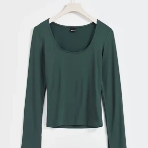 Mörkgrön långärmad topp - En stilren mörkgrön långärmad topp från ginatricot med rundad, djup urringning. Modellen är croppad och har en slim passform som sitter snyggt på kroppen. Perfekt att matcha med jeans eller kjol för en clean look.