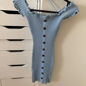 Ljusblå ribbad kortklänning Mooloola - Supersöt ljusblå ribbad kortklänning från Mooloola med offshoulder och volangkant. Klänningen har bruna knappar hela vägen framtill och är figurnära med stretchigt material. Perfekt för dig som gillar en feminin och trendig look! Klänningen har en liten tygskavank som knappt syns (se bild).