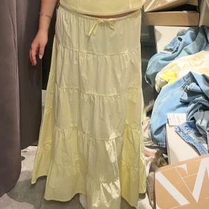 Ljusgul volangkjol med knyt - Supersöt långkjol i ljusgul färg med flera volanger och knyt i midjan. Kjolen har en luftig och flowy känsla, perfekt för varma dagar. Passar dig som gillar en romantisk och avslappnad stil.