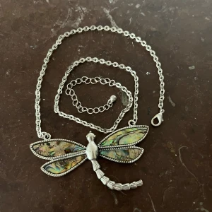 trollslända halsband - Unikt halsband med en stor trollslända som hänge. Vingarna har skimrande i , färgglada inslag i grönt, blått och gult, inramade av silverfärgad metall. Kedjan är justerbar och har ett klassiskt lås. Perfekt för dig som gillar naturinspirerade smycken. på sista bulden kan nu se storleken jämfört med ett gem