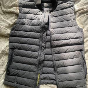 Svart dunväst från J.Lindeberg - Snygg svart dunväst från J.Lindeberg med quiltad design och dragkedja framtill. Västen har två fickor med dragkedja och en gul J.Lindeberg-logga på ryggen.  Endast använd 1 gång! Nypris 1200.