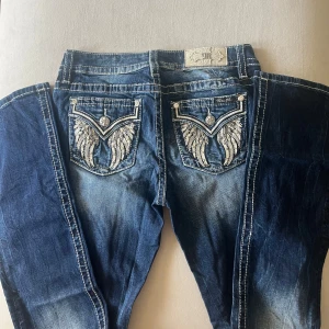 Miss Me low bootcut jeans med brodyr - Snygga blå Miss Me low bootcut jeans med coola broderade fickor med vingar bak. Tydliga kontrastsömmar och klassisk bootcut passform. Jeansen har låg midja och är gjorda i stretchig bomullsdenim för en bekväm känsla. Innerbenslängd: 84cm Midjevidd: 37,5cm Totallängd: 112,5cm Benvidd nedtill: 23cm