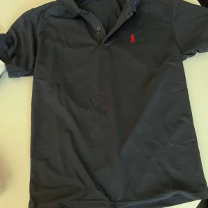 Svart pikétröja från Polo Ralph Lauren - Klassisk svart pikétröja från Polo Ralph Lauren med röd broderad logga på bröstet. Tröjan har krage, två knappar vid halsen och är tillverkad i mjuk bomull. Perfekt för en clean och stilren look.