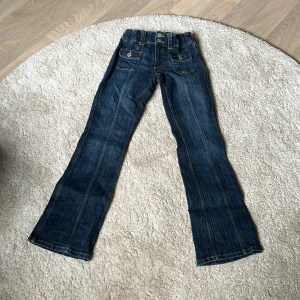 Bootcut jeans från H&M, mörkblå - Mörkblå bootcut jeans från H&M med låg midja och justerbar resår. Klassisk femficksmodell med två fickor fram med lock och knappar. Snygga kontrastsömmar och slitstarka i jeansmaterial. Perfekta för en cool och avslappnad stil.