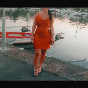 Orange kort klänning med bälte - Snygg orange kortklänning med smala axelband och omlott-detalj i midjan med bälte. Klänningen har en figurnära passform och är perfekt för varma dagar. Materialet ser ut att vara mjukt och lätt, vilket ger en skön känsla.