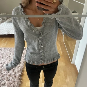 Grå kofta med volang och knappar - Supersöt grå kofta med volangkant längs knäppningen och nederkanten. Långärmad och stickad i mjukt material, perfekt för lager-på-lager. Knappar framtill och feminin touch med volanger. Passar dig som gillar en söt och chill stil.