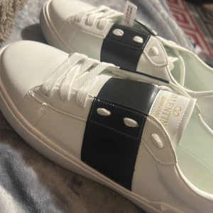 Valentino Garavani Open sneakers vit/svart - Snygga Valentino Garavani Open sneakers i vitt skinn med bred svart läderdetalj över mitten och ikoniska nitar baktill. Klassisk rund tå, vita snören och guldlogga på plösen. Perfekta för dig som vill ha en clean men lyxig look.