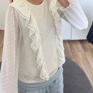 Vit blus med volang från Gina Tricot - Supersöt vit blus från Gina Tricot med diskret prickigt mönster och stora volanger framtill. Långärmad modell i lätt och luftig bomullsmix, perfekt för vår och sommar. Stängs med knapp i nacken. Ger en romantisk och trendig vibe!