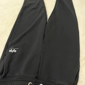 Svarta mjukisbyxor Alpha Industries - Svarta mjukisbyxor från Alpha Industries med logga på benet. Byxorna har snörning i midjan och är tillverkade i mjukt bomullsmaterial. Perfekta för en avslappnad och chill stil.