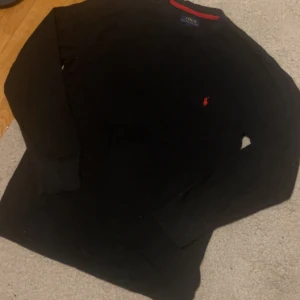 Svart långärmad tröja Polo Ralph Lauren - Svart långärmad tröja från Polo Ralph Lauren med klassisk röd broderad logga på bröstet. Tröjan har rund halsringning med röd detalj på insidan och är tillverkad i mjuk bomull. Perfekt för dig som gillar stilren och enkel design.  Kommer strykas och tvättas efter köp 