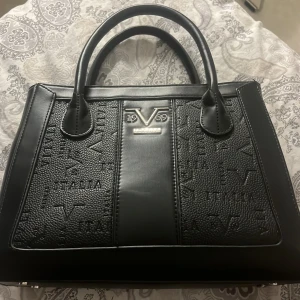 Svart handväska från V1969 Italia - Snygg svart handväska från V1969 Italia med präglad logga och text på sidorna. Väskan har två rejäla handtag, 5 fack med dragkedja och detaljer i silver. Tillverkad i skinnimitation med stilren, boxig form och struktur på ytan. Fint skick☺️Köpt för 1700kr😊