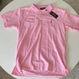 Rosa pikétröja från Polo Ralph Lauren - Snygg rosa pikétröja från Polo Ralph Lauren i bomull. Klassisk krage, korta ärmar och blå broderad logga på bröstet. Modellen är slim fit och passar perfekt för en clean och fräsch stil.