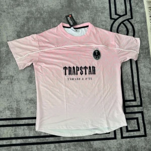 Rosa Trapstar fotbollströja  - Snygg rosa fotbollströja från Trapstar med svart logga och texten 'IT'S A SECRET' på bröstet. Tröjan har korta ärmar, vit piping och ett stort nummer 22 på ryggen. Tillverkad i lätt och ventilerande meshmaterial, perfekt för dig som gillar streetwear och sportig stil.