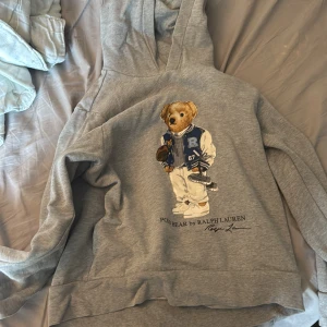 Grå hoodie Polo Bear Ralph Lauren - Grå hoodie från Polo Ralph Lauren med ikoniska Polo Bear-trycket framtill. Tröjan har huva, långa ärmar och en avslappnad passform. Perfekt för dig som gillar streetstyle och klassiska märken liten i storleken men fråga på.