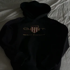 Svart hoodie från GANT med tryck - Svart hoodie från GANT med stort broderat logotyp-tryck och sköld på bröstet. Klassisk huva och ribbade muddar. Snygg och sportig stil, perfekt för dig som gillar streetwear och vill ha något bekvämt och trendigt.