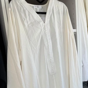 Vit modest Abaya  - Vit/creme vit abaya med v-ringning och dekorativa knappar längs bröstet från jeansindustry. Abayan har långa ärmar och fina broderade detaljer runt halsringningen och längs knäppningen. Band/skärp medföljer. Luftigt material som ger en elegant outfit💕 Storlek M/L. Lappar kvar
