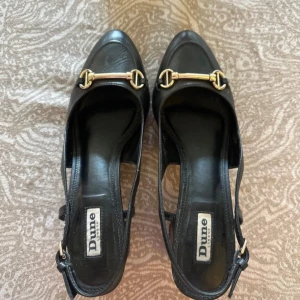 Svarta klackskor från Dune London - Stilrena svarta klackskor i skinn från Dune London med guldfärgade metalldetaljer framtill. Skorna har en spetsig tå, öppen häl med justerbart spänne och en stabil låg klack. Perfekta för dig som gillar klassisk och elegant stil.