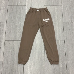 Bruna mjukisbyxor från Shein - Bruna mjukisbyxor från Shein med tryck 'New York' på ena benet. Byxorna har resår i midjan med vit snörning, sidofickor och muddar vid bensluten. Perfekta för chill dagar eller när du vill vara bekväm.