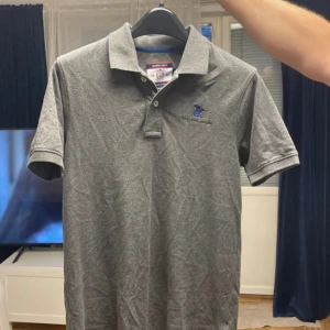 Grå pikétröja Beverly Hills Polo Club i underbar skick - Snygg grå pikétröja från Beverly Hills Polo Club med klassisk krage och knappar framtill. Broderad blå logga på bröstet ger en sportig vibe. Skön passform och tillverkad i mjuk bomull, perfekt till jeans eller shorts. Köpt från Dubai Beverly hills polo club butik för ung 900kr