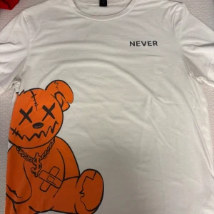 Vit t-shirt med orange björntryck - Vit t-shirt med coolt tryck av en orange tecknad björn med korsade ögon och kedja, samt texten 'NEVER' på bröstet. Klassisk passform och rund hals. Perfekt för dig som gillar streetstyle och unika motiv.